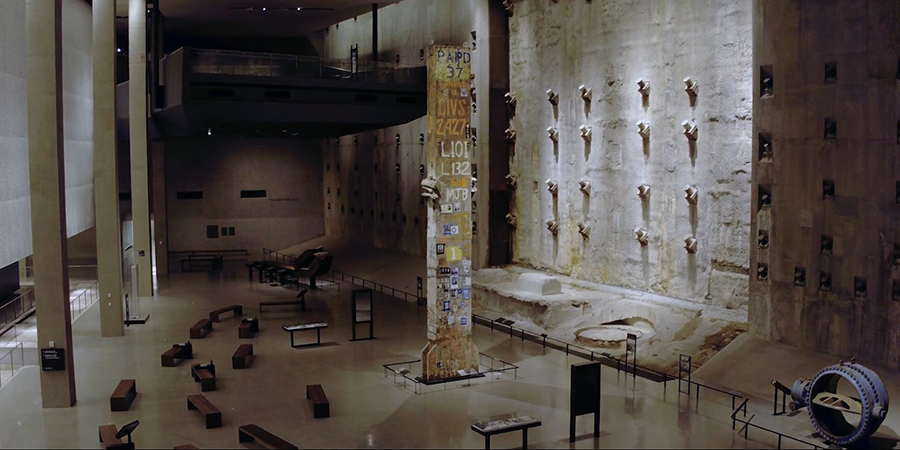 This view from a drone captures Foundation Hall from above. The Last Column is standing in the center of the hall and is surrounded by wooden benches. The slurry wall is lit up to the right. Shadowed areas of the Museum are in the background.