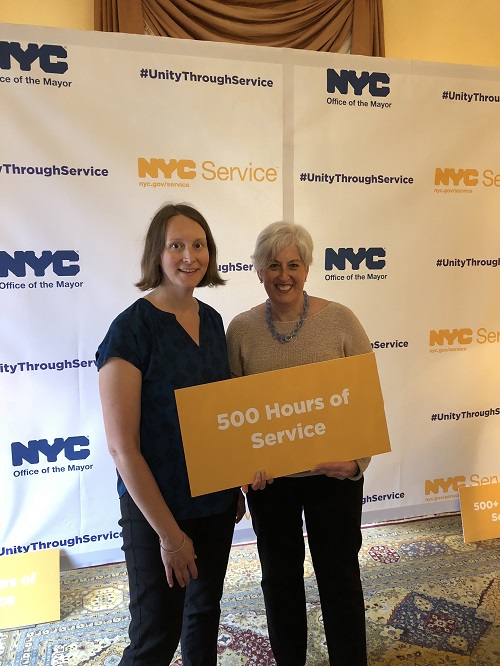 Karen Lazar, a visitor services volunteer and docent, holds a sign that reads “500 hours of service” while visiting Gracie Mansion for the 2017 Mayoral Service Recognition Award ceremony. She is standing with Lindsay Watts, director of the 9/11 Memorial & Museum’s volunteer program.