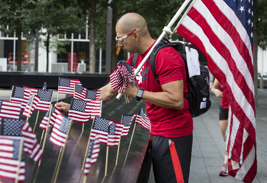 A volunteer with Team Red, White & Blue helps place 2,983 flags at the Memorial. He is holding a bunch of small flags in his left hand as he places each flag with his right hand. He is also holding a large American flag over his left shoulder.