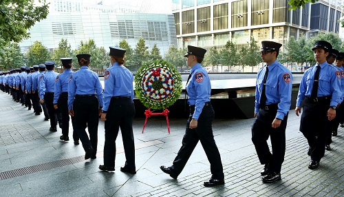 Dozens of formally dressed FDNY probationary EMTs and medics gather along the south pool for a wreath laying ceremony to honor fire department members who lost their lives on 9/11. A large wreath stands in front of the south pool as they walk by.