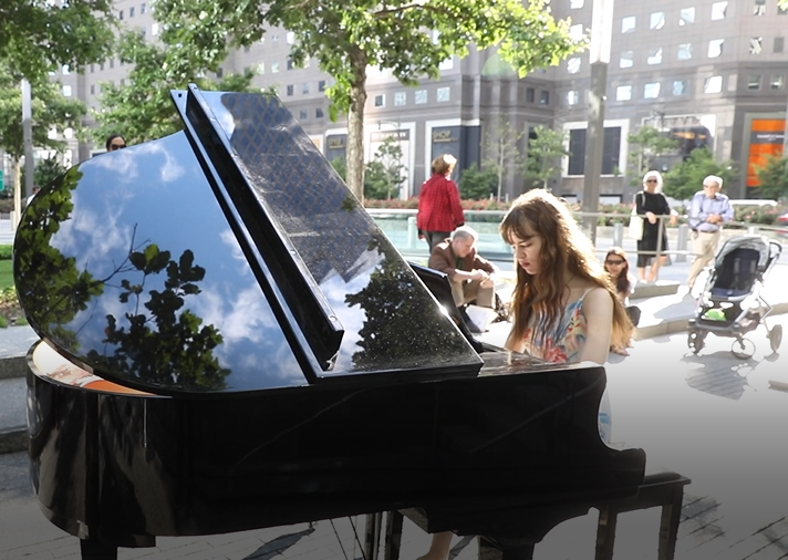A young woman plays a black piano on the Memorial plaza as part of Make Music New York. Several people watch on in the background.