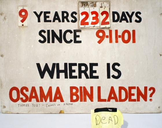 A sign that tracked the days Osama bin Laden was at large is displayed on a white surface. The sign reads: “9 years, 232 days since 9/11/01. Where is Osama bin Laden?” A yellow piece of paper beside the sign reads “Dead.”