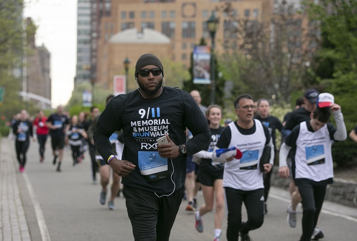 A man with a black hat, sunglasses, and black running outfit leads a pack of runners on an overcast day.