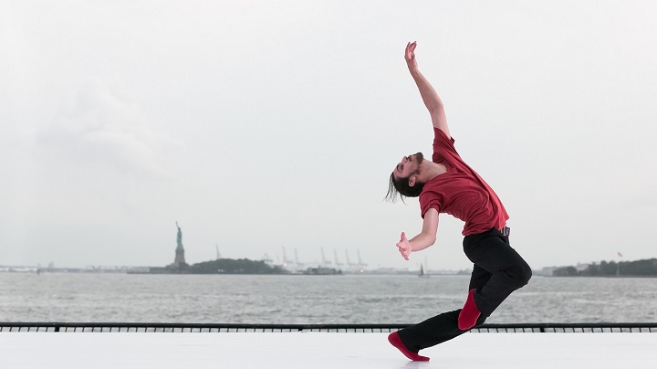 A ballerina dancer in red shirt and black leggings performs on a white platform. The Statue of Liberty is visible in the distance.