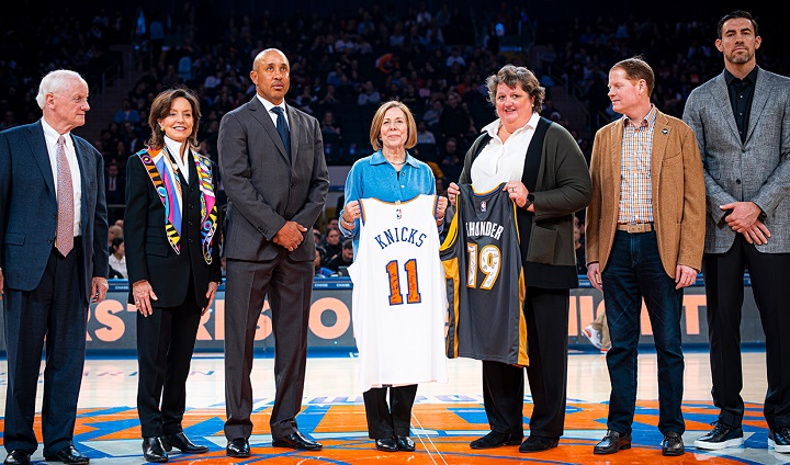 Seven people on a basketball court. In the center, two women stand holding basketball jerseys for New York Knicks and the Oklahoma City Thunder, respectively. All seven are standing in a row and looking into the camera.