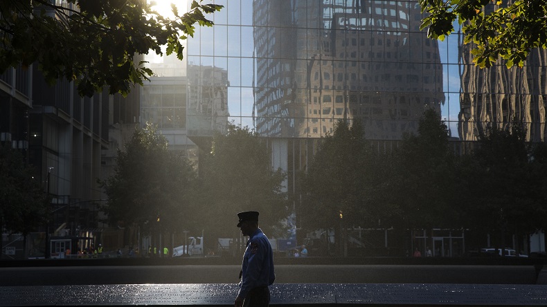 A firefighter in a formal outfit and hat looks at victims’ names on a bronze parapet. Rays of sunlight come through a gap in buildings and shine down on him and a reflecting pool. In the darkened distance are trees and building facades.
