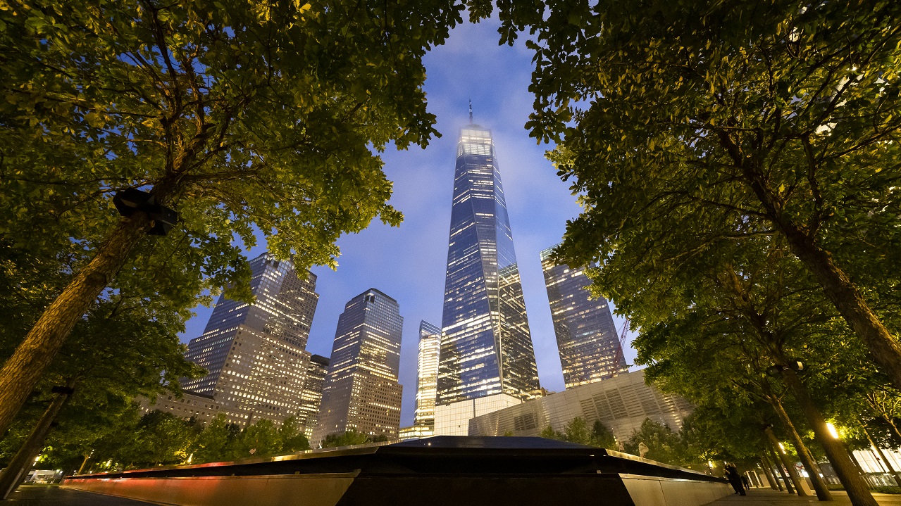 One World Trade Center appears through a parting of the trees on the 9/11 Memorial plaza at dusk.