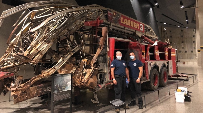 Two firefighters in masks and navy blue T-shirts stand in front of the smashed Ladder 3 firetruck in the 9/11 Memorial Museum.
