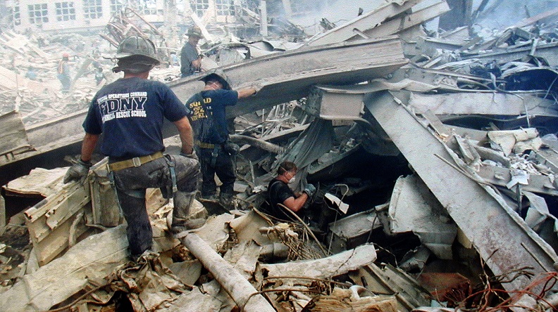 Two firefighters in masks and navy blue T-shirts wade through the rubble and twisted steel at Ground Zero while another descends into a cravasse. 