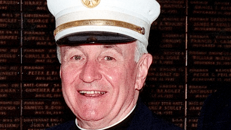Father Mychal Judge, an FDNY chaplain, smiles brightly for a photo in a formal FDNY outfit including a gold-trimmed white cap with a navy brim..
