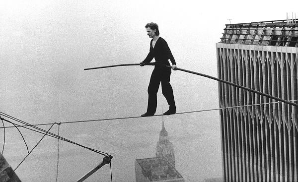 A black and white historical photo shows Philippe Petit holding a long pole as he does a hire-wire walk between the Twin Towers. The buildings of lower Manhattan can be seen below him.