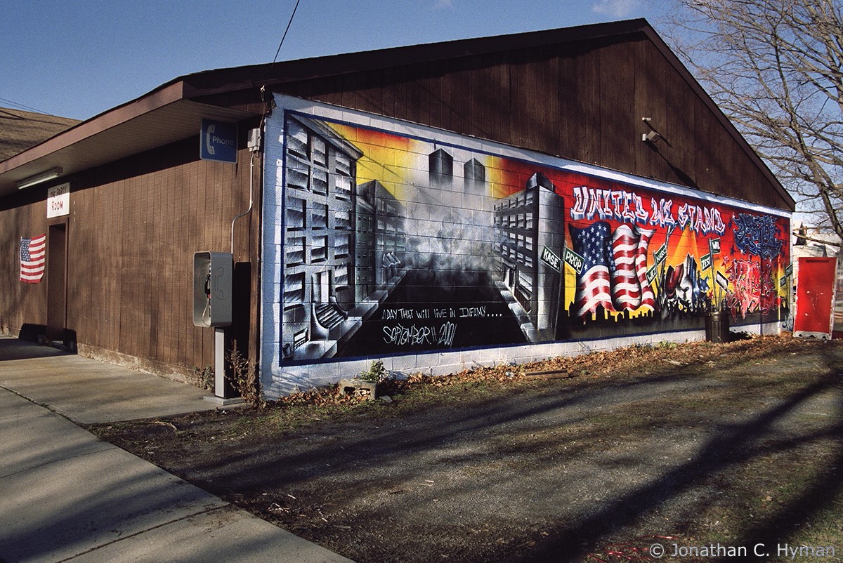 Graffiti mural depicting the Twin Towers on a smoke-filled city block with the text "A Day That Will Live in Infamy." On the other side, a giant American flag amid city street signs and the text "United We Stand."