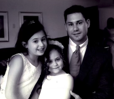 Two little girls in white dresses smile while sitting with their father, in a dark suit