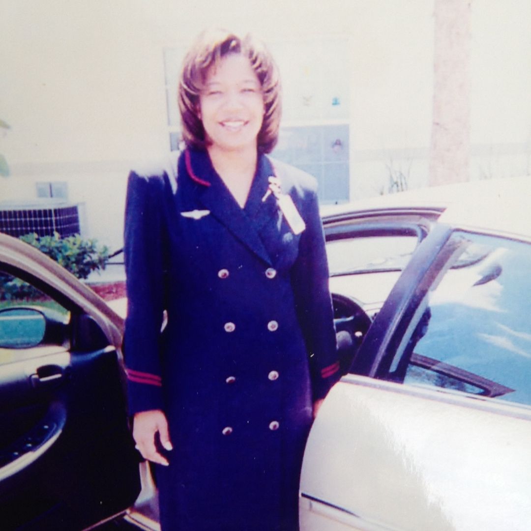 A dark-haired woman in a navy blue flight attendant uniform smiles in front of a white car