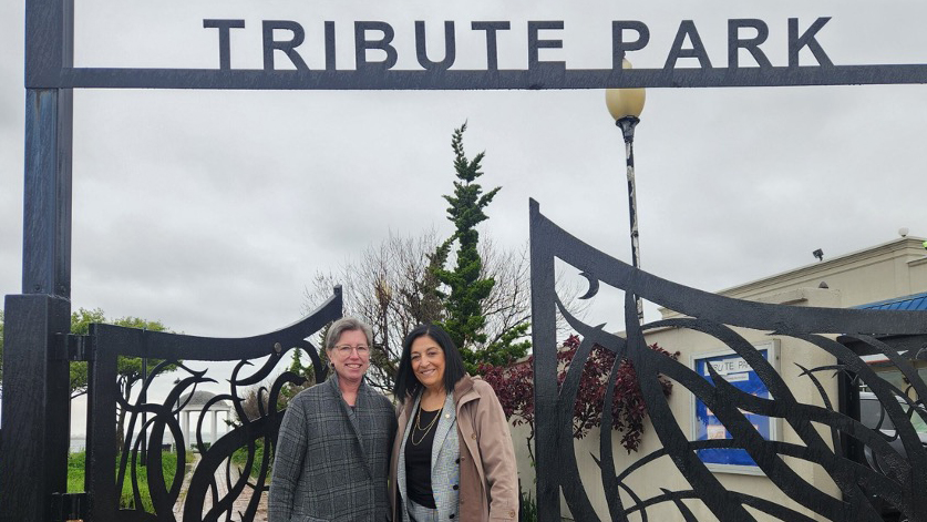Elizabeth Hillman and  Joann Ariola pose in front of a decorative black metal gate that says Tribute Park