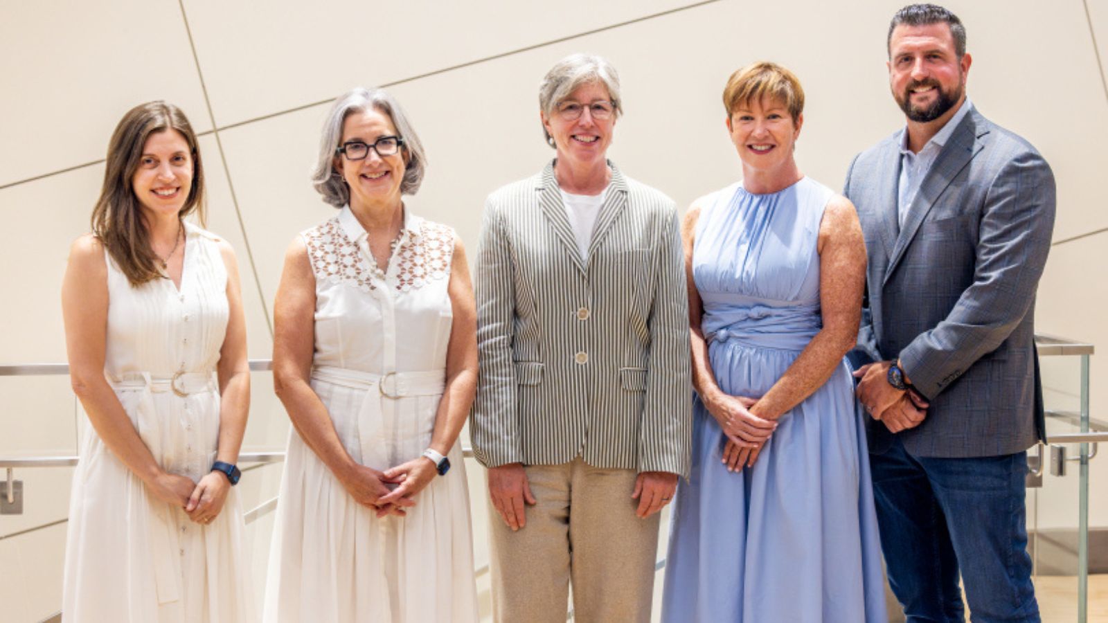 A group of five faces the camera, smiling and standing in front of an off-white background.