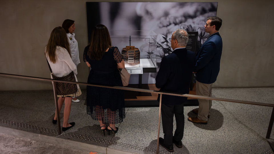 Five formally dressed adults stand, backs to camera, looking at a wooden octagonal memorial stand covered with engraved black and brass plaques.