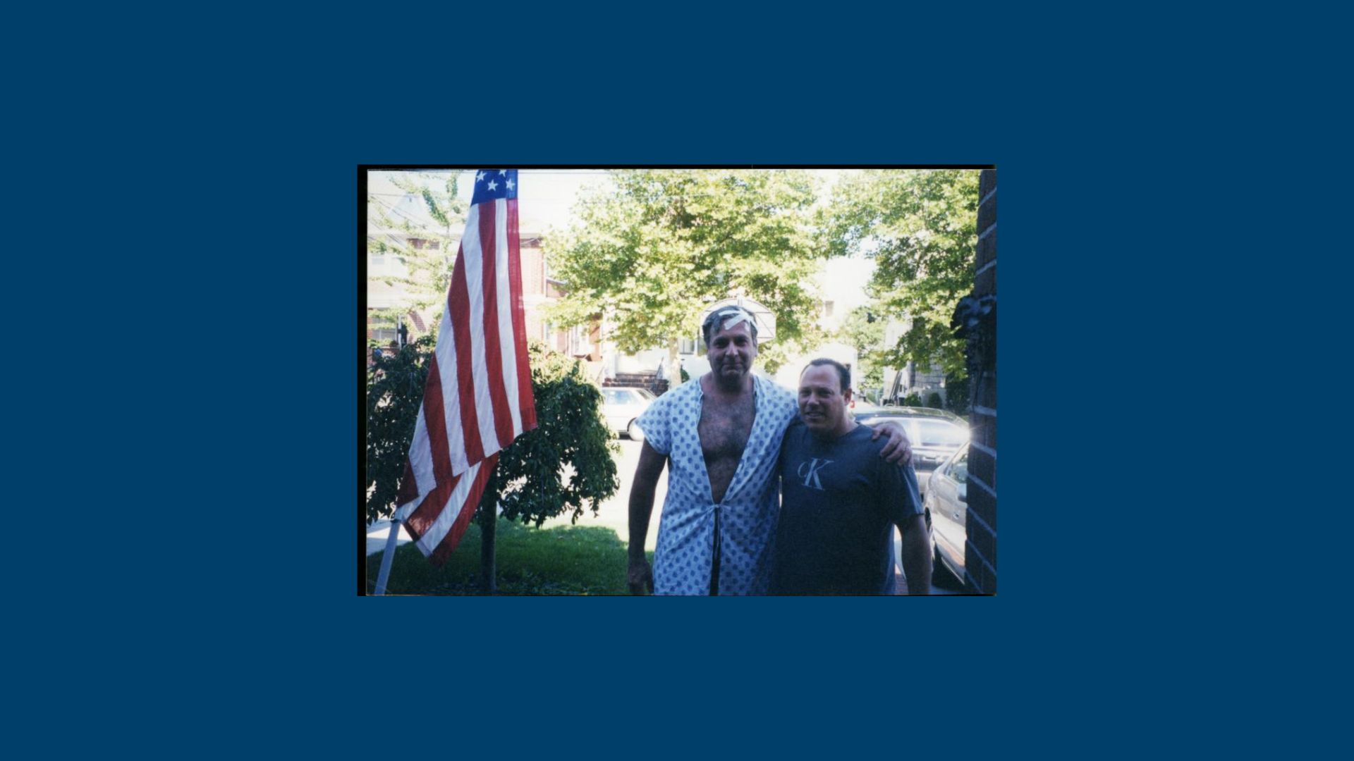Mr. Eichen stands in a hospital gown with the man who took him home. They are beside an American flag.