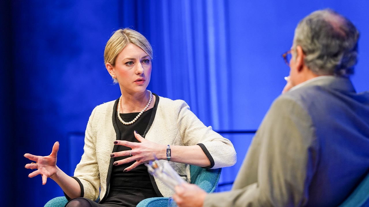 In this close-up view, author Joana Cook gestures with both hands as she speaks with moderator Clifford Chanin onstage at the Museum Auditorium. Chanin is in the foreground and out of focus. A wall behind the two of them is lit blue.