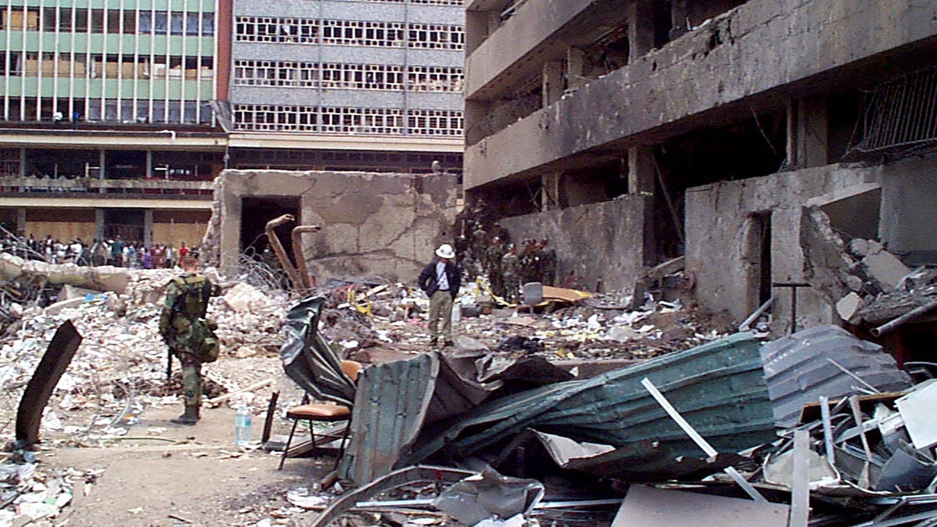 groups of civilians, soldiers in camouflage, and a person in a white hat standing among the debris next to a bombed building