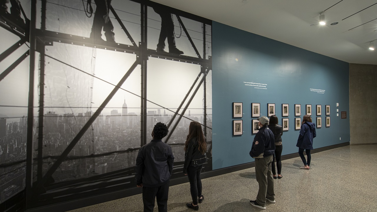 Men and women looks at a large black-and-white photograph and 16 tintype photographs displayed on a blue wall, which is part of the special exhibition “Skywalkers: A Portrait of the Mohawk Ironworker at the World Trade Center.”