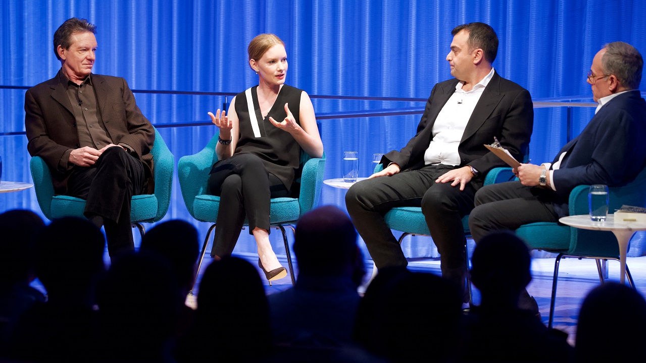 Three men and a woman take part in a moderated discussion on stage at the Museum. The woman is speaking, second from the left, as the three men listen. The black silhouettes of audience members are in the foreground.