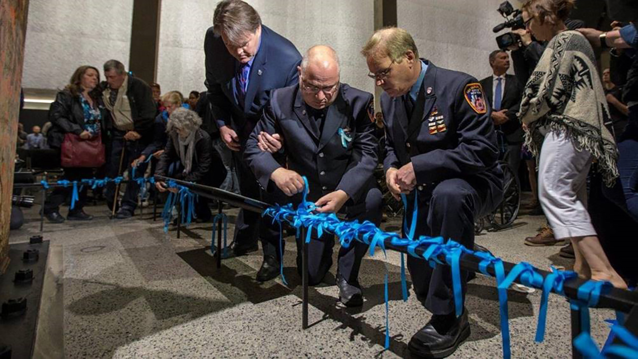 Several members of the FDNY and the family members place blue ribbons on a black railing at the foot of the Last Column.
