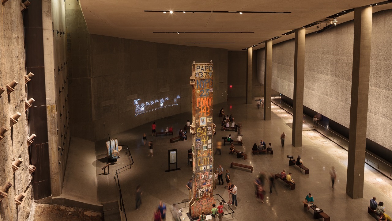A view from above shows visitors gathering around the Last Column, a thirty-six-foot-tall steel beam that was the last to be removed from Ground Zero. In the distance, visitors observe information about 9/11 projected on a wall.