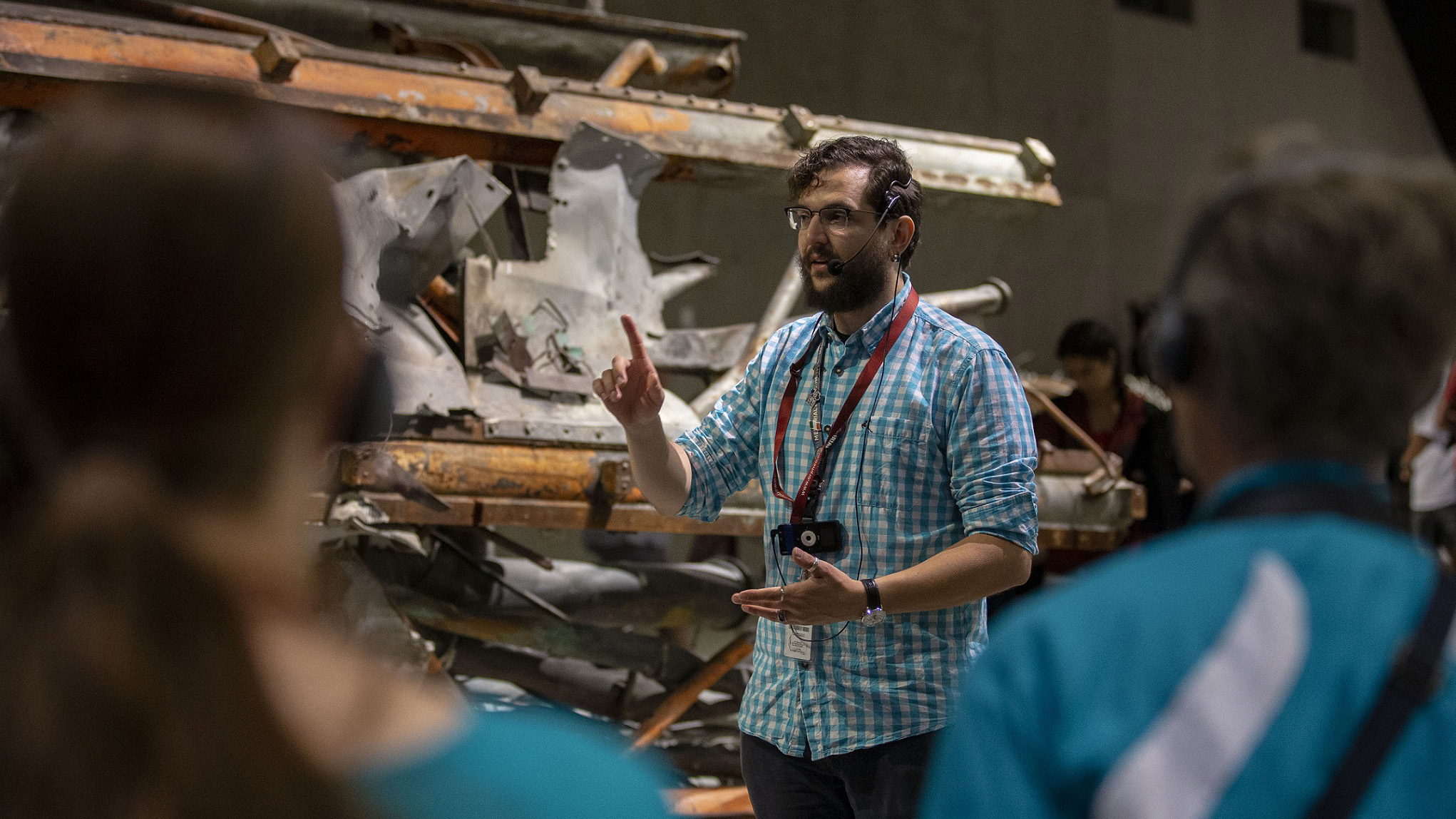 A bearded young male tour guide, standing in front of a mangled segment of an antenna recovered from the World Trade Center, gestures as talks to visitors on a tour of the 9/11 Memorial Museum.