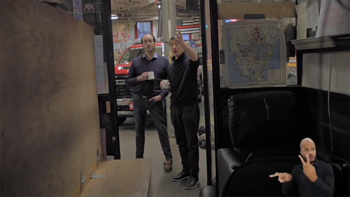 Two men standing in an inner doorway of a fire station, holding cups of coffee and talking. Behind them are a fire truck and an American flag with red firefighter stripe. An ASL interpreter is present in the lower right corner of the screen.