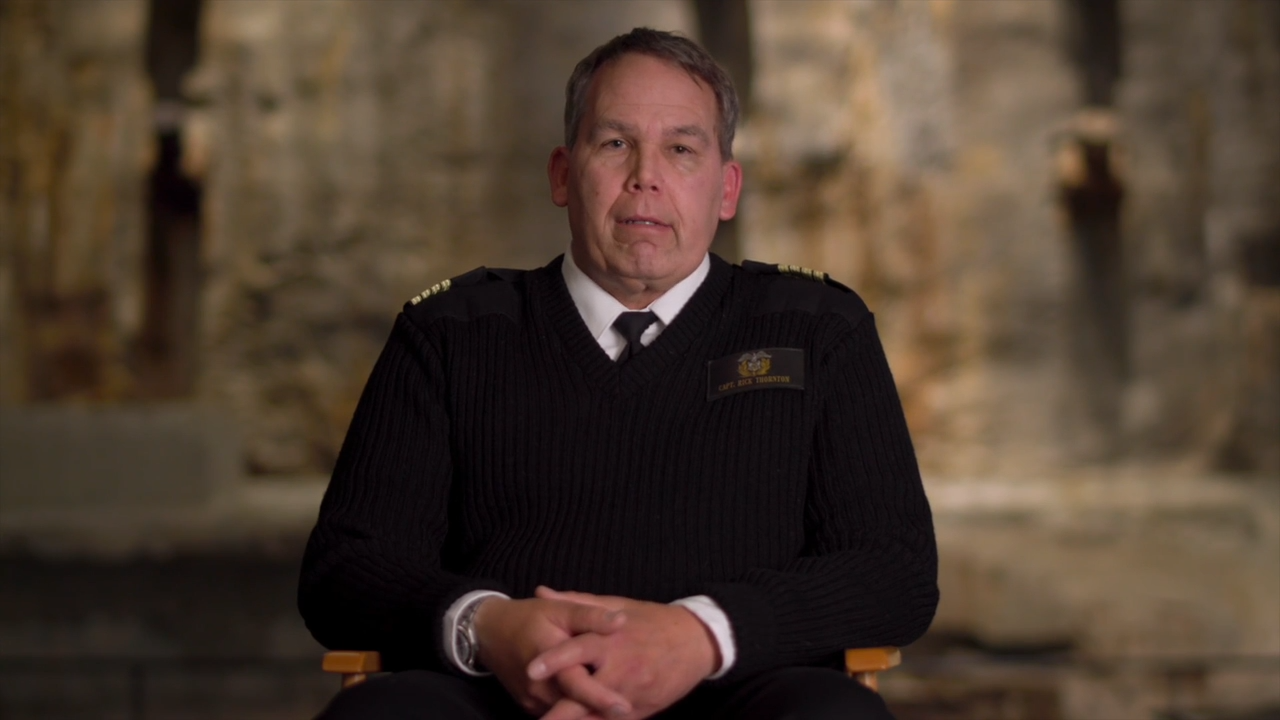 A man wearing a black uniform with gold epaulets sits with hands folded.
