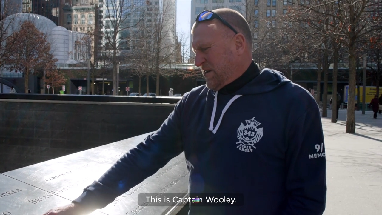 Tim Brown has short cropped hair and wears an FDNY sweatshirt. He stands at the 9/11 Memorial, where victims' names are engraved on waist-high walls.
