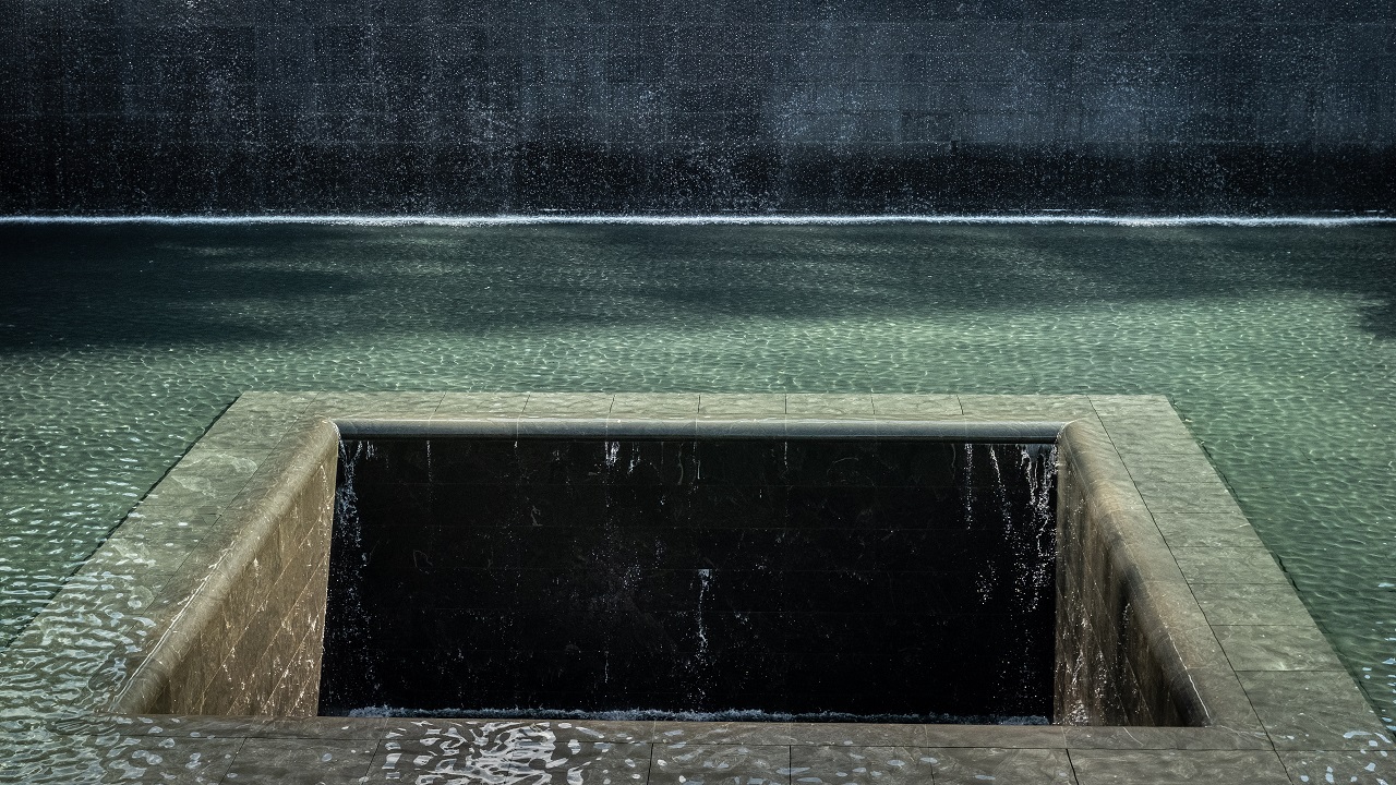 Water flows down four walls and disappears into a void at the center of a Memorial reflecting pool. Sunlight creates shadows on the pooled water.