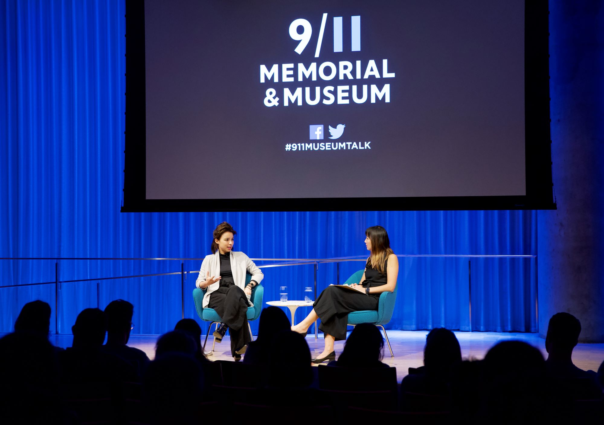 VICE correspondent Isobel Yeung speaks onstage as she sits with a woman hosting a public program at the Museum Auditorium. This wide-angle photo includes audience members in the foreground who are silhouetted by the light onstage. A projector screen has been lowered above the two women onstage.