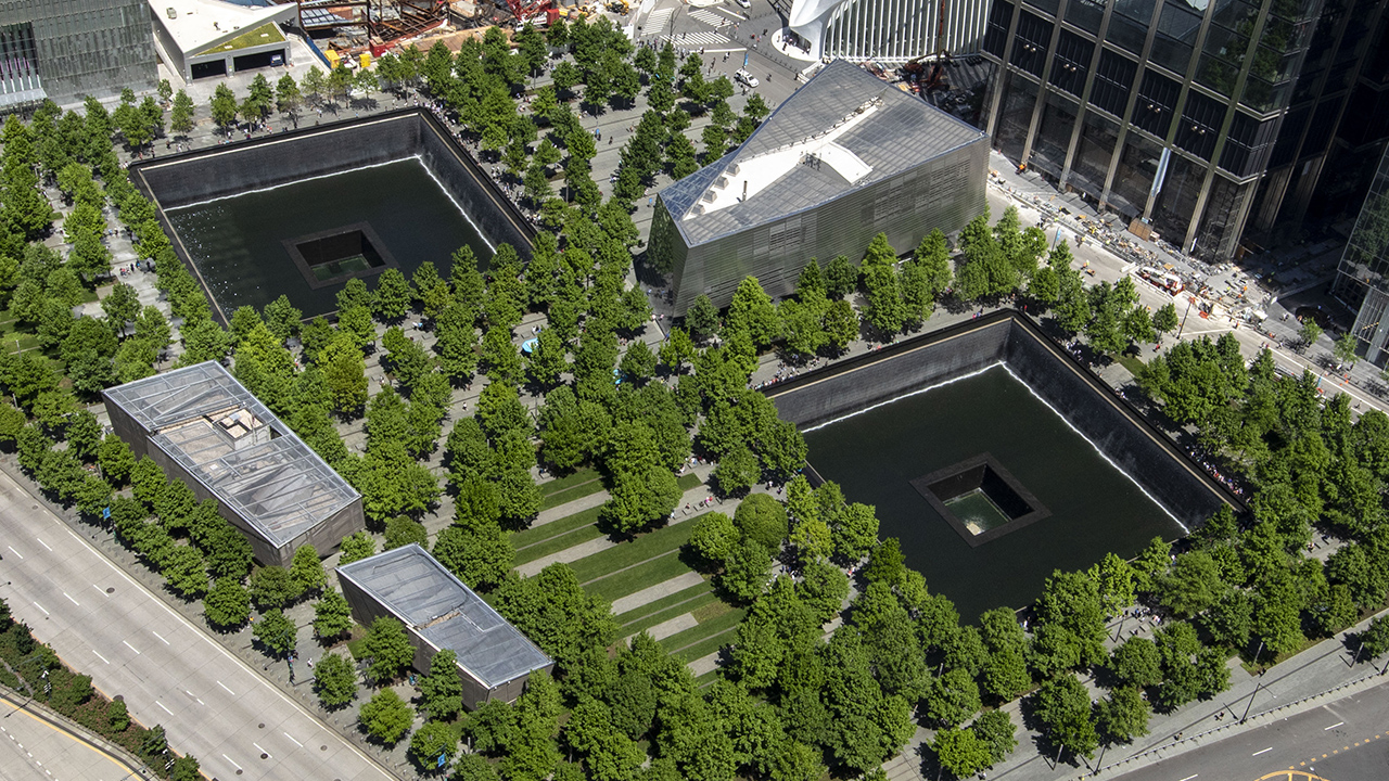 Memorial Plaza is seen from above on a sunny day. Dozens of oak trees fill the Plaza with a vivid green. These trees surround the two reflecting pools where the North and South towers once stood. Between the large, square pools is the Museum’s glass and steel pavilion.