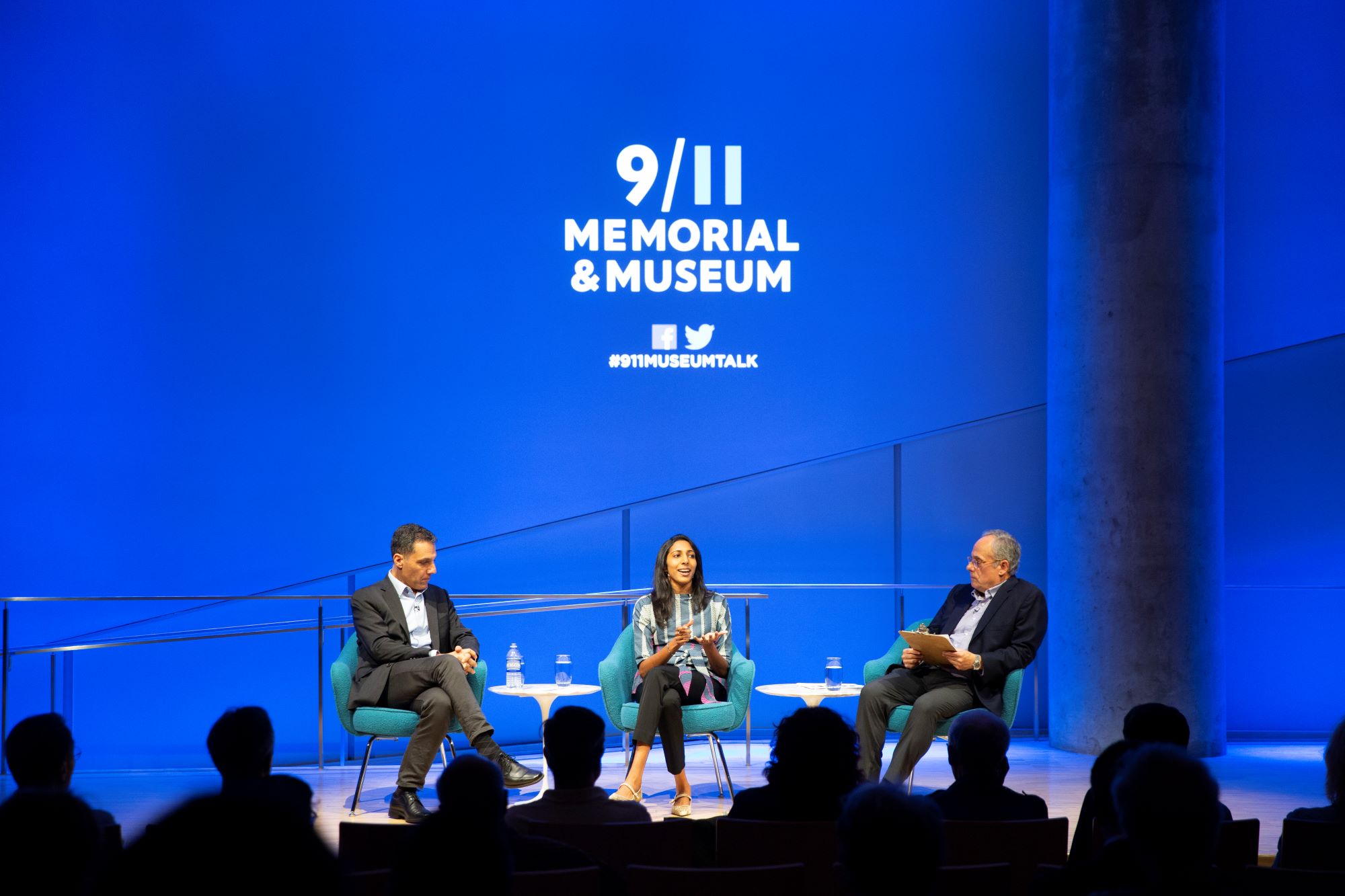 Hany Farid, Dartmouth computer science professor and senior advisor to the Counter Extremism Project, and Vidhya Ramalingam, founder of Moonshot CVE, sit onstage during a public program in the Museum Auditorium. Clifford Chanin, the executive vice president and deputy director for museum programs, sits to the left of them holding a clipboard. Blue and white lights illuminate the stage and silhouette audience members in the foreground. The 9/11 Memorial & Museum logo is projected above them.