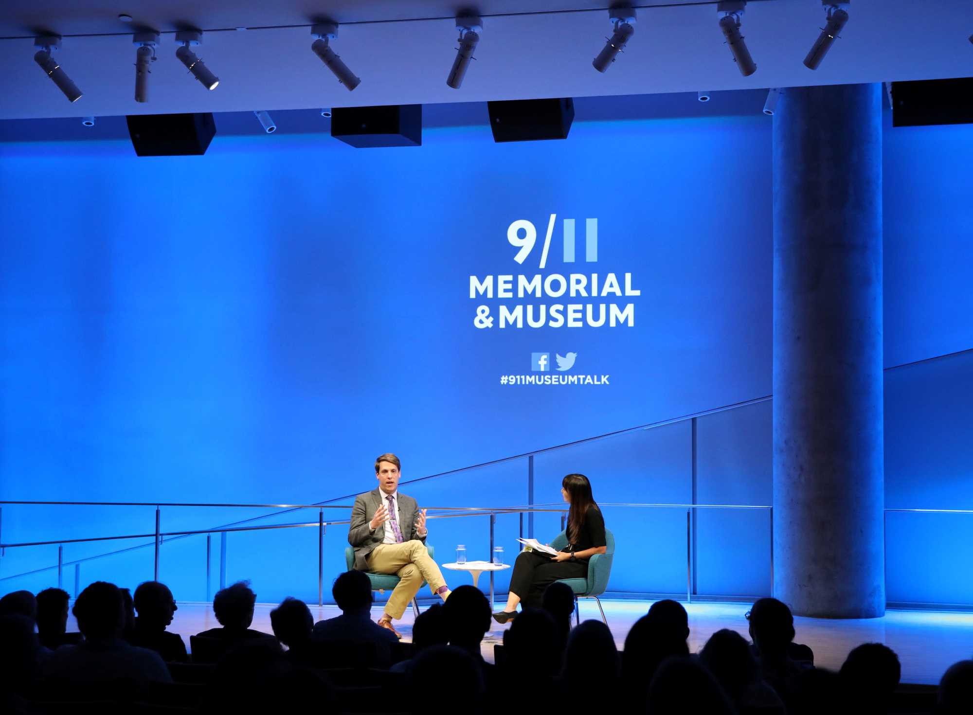 A wide shot of the Museum Auditorium shows journalist and historian Garrett Graff gesturing with both hands as he speaks onstage at the public program, Only Plane in the Sky. A woman sitting next to him who is hosting the event listens while holding a clipboard. Audience members are silhouetted in the foreground by blue light behind onstage.