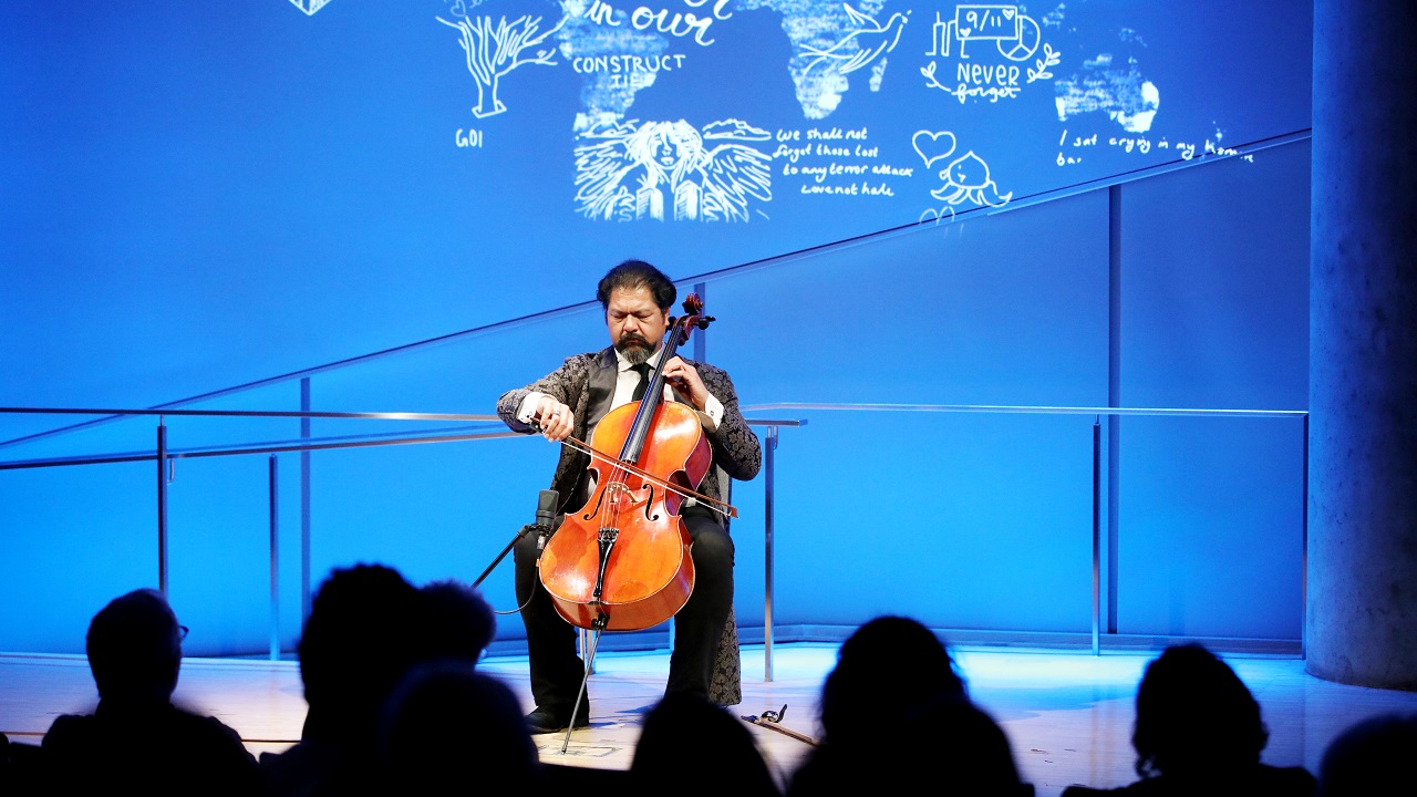Cellist Karim Wasfi performs while seated onstage in this wide-angle photo of the Museum Auditorium. His bright orange-brown cello contrasts with the blue lights of the stage. The silhouettes of audience members are visible in the foreground.