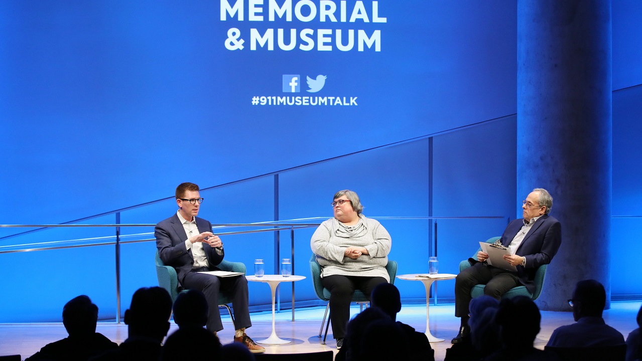 David Tessler of Facebook speaks onstage as Joan Donovan of the Harvard Kennedy School and Clifford Chanin, the executive vice president and deputy director for museum programs, look on to his left. The silhouettes of audience members are visible in the foreground. The wall behind them is lit up blue and the logo of the 9/11 Memorial & Museum is displayed on it.