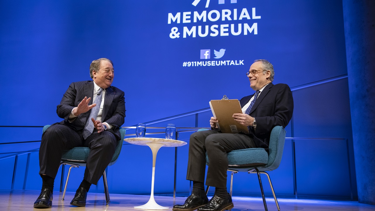 Real estate owner and builder Howard Milstein gestures with his left hand as he sits on the Museum Auditorium stage with moderator Clifford Chanin, executive vice president and deputy director for Museum Programs. Milstein and Chanin are both smiling as they sit on the stage, which is bathed in blue and white light. A white table with two glasses of water sits between the two of them.