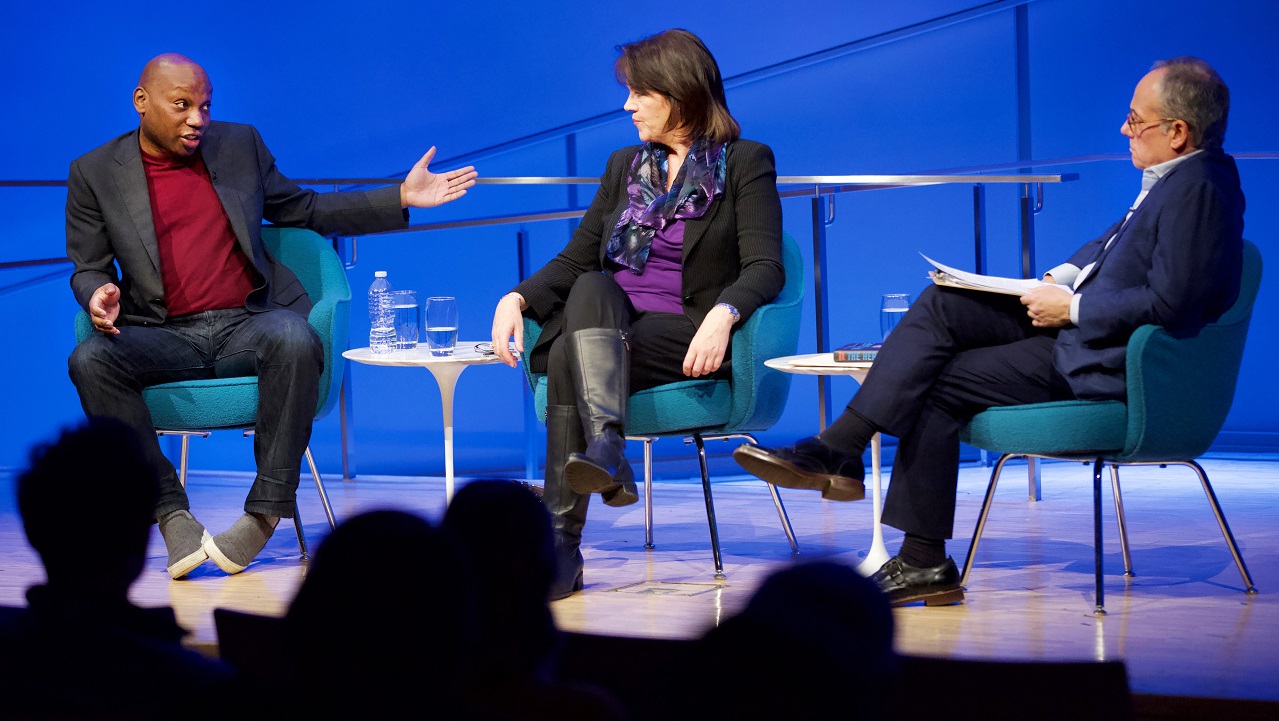 Three public program participants sit on a blue-lit auditorium stage. To the far left, a man in a gray blazer, red sweater, and jeans gestures with both arms, his face holds an expression of incredulity. A woman in the center and the moderator look on. The heads of the audience members appear in the foreground in silhouette.
