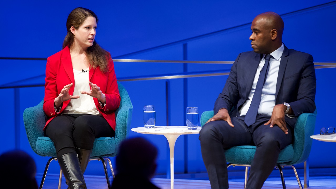 In this close-cropped photograph, two public program participants sit on a blue-lit auditorium stage. To the left, a woman in a red blazer gestures with her hands while the other participant, a man in a suit, looks in her direction.