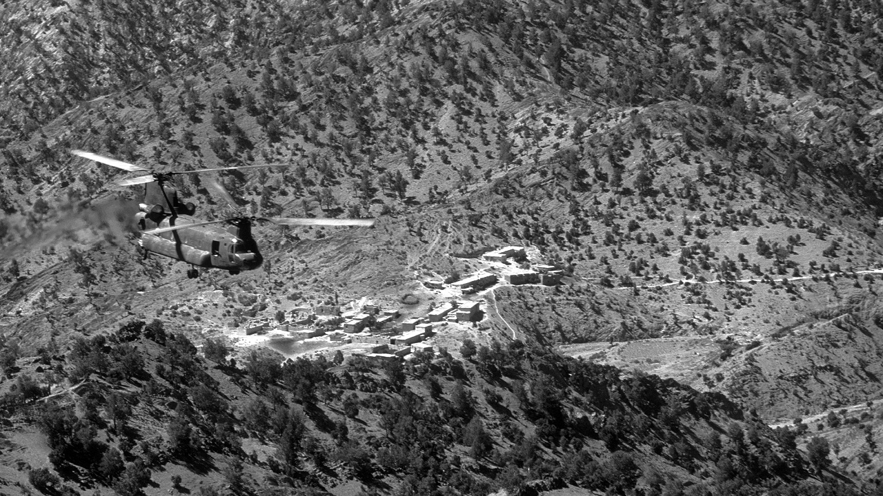 Birds eye view of a tandem rotor helicopter flying over a rocky mountain terrain.
