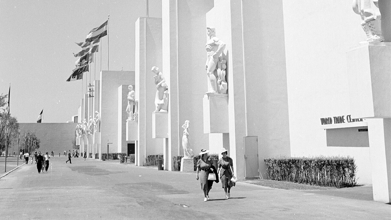 Black and white photograph of an angled pathway with two indiscernible women walking along a white building. Flags are visible on the building in the background.
