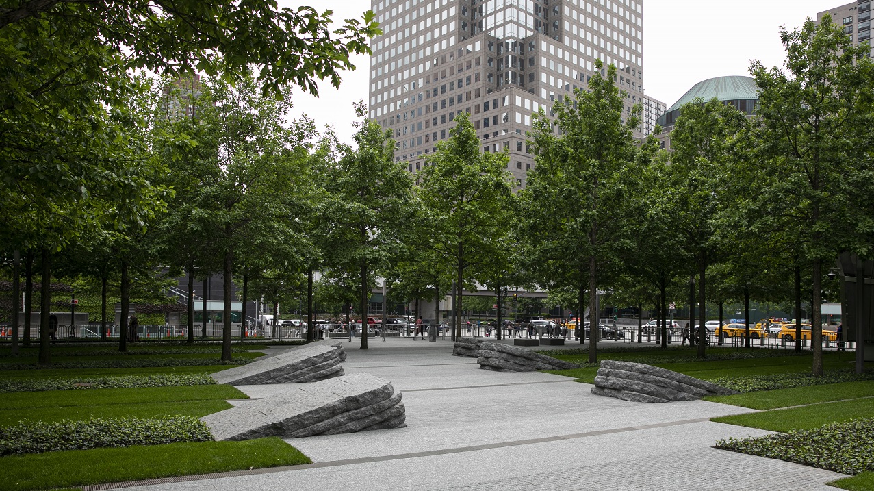 This photograph shows the 9/11 Memorial Glade on an overcast day, shaded by a canopy of trees with green leaves.