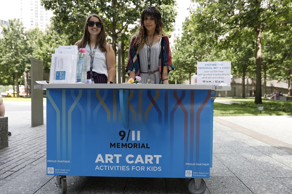 Two brown-haired women stand on the plaza behind a blue cart with a white top