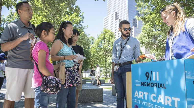 A family of four - a man, woman, young girl, and young boy - looks on as two Museum staffers stand at the Art Cart on the plaza. Green foliage is visible in the background.