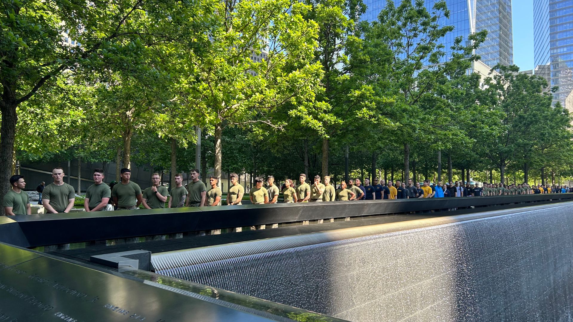 A line of military members in army green, navy blue, and bright yellow tee shirts lines one side of the Memorial pool. Behind them is a row of lush green trees in sunlight, and the corner of One World Trade is viewable at top right.