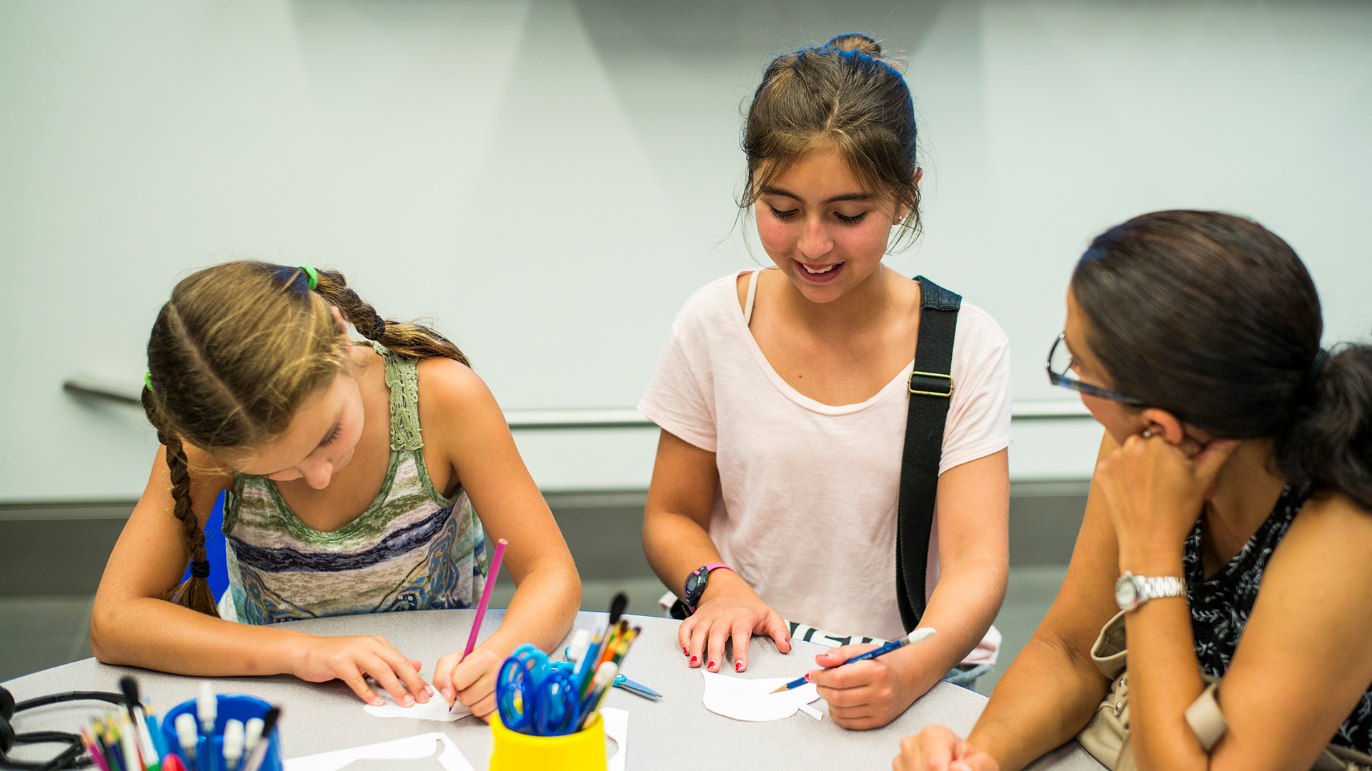 Two children color at a table while an adult looks on