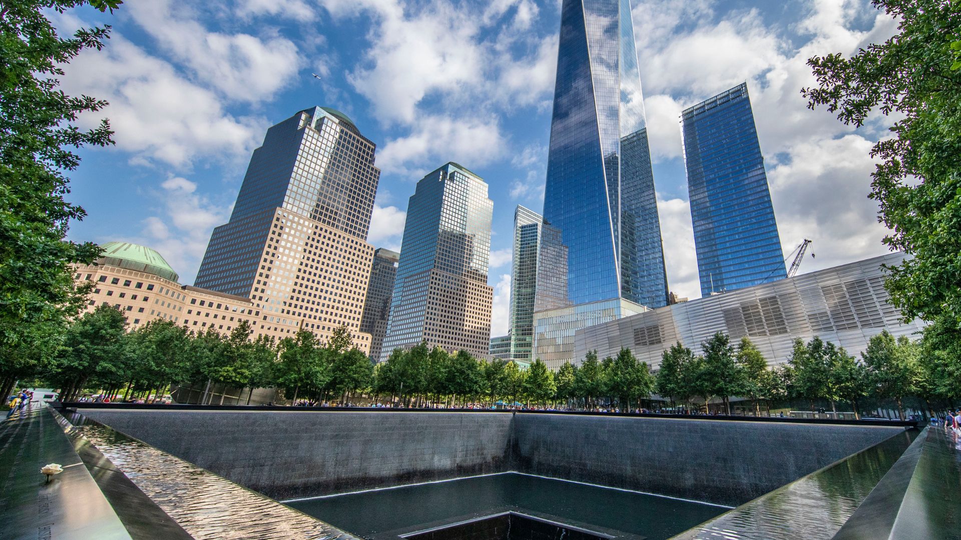 One of the Memorial reflecting pools and its parapets, surrounded by green trees and a portion of the skyline including One World Trade.
